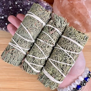 May include: Three bundles of dried green sage, wrapped with white twine. The sage bundles are displayed against a wooden surface, with a purple crystal and a pink crystal in the background. A bracelet with white and blue beads is visible.