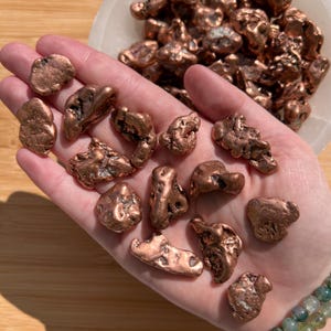 May include: A hand holding several raw copper nuggets with a rough, textured surface. The nuggets vary in size and shape, displaying a warm, reddish-brown color. More copper nuggets are in a white bowl in the background.