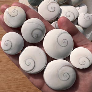 Pode incluir: Uma coleção de pedras brancas e redondas com um desenho em espiral cinza. As pedras têm uma superfície lisa e polida. O padrão em espiral é de um tom cinza mais escuro, criando contraste. As pedras são seguradas em uma mão e variam em tamanho.