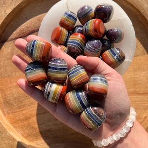 Op de afbeelding: Een verzameling getrommelde regenboogobsidiaanstenen, elk met meerdere lagen kleur, waaronder rood, oranje, geel, groen, blauw en paars. De stenen zijn in een hand gerangschikt, waardoor hun unieke patronen en texturen worden getoond.