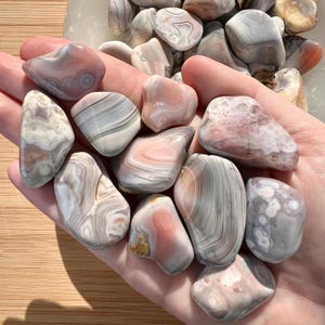 May include: A collection of polished agate stones in various shapes and sizes. The stones display a mix of colors, including pink, gray, and white, with banded patterns and swirls. Some stones have a translucent quality.