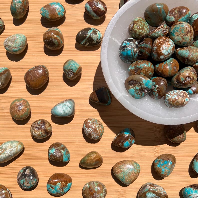 Pu&ograve; includere: Una collezione di pietre di turchese levigate e arrotondate con venature di matrice marrone sparse su una superficie, con un folto gruppo di queste pietre contenute all'interno di una ciotola circolare bianca e traslucida con una finitura opaca liscia.