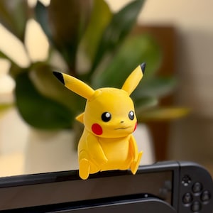 May include: A yellow Pikachu figurine with black-tipped ears and red cheek circles. The figurine is perched on a black electronic device, with a blurred green plant in the background. The Pikachu figure is a popular character from the Pokemon franchise.