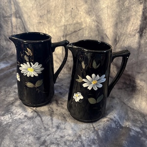 May include: Two black ceramic pitchers with white daisy floral designs. Each pitcher has a handle and a spout. The daisies have yellow centers and green leaves. The pitchers are likely for serving beverages.
