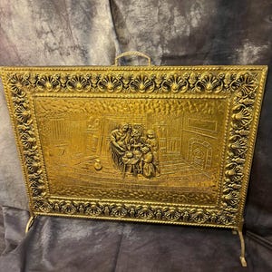 May include: A gold-colored fireplace screen with a detailed relief scene of people gathered around a table. The screen has a decorative border and a handle at the top. The screen is supported by two legs.