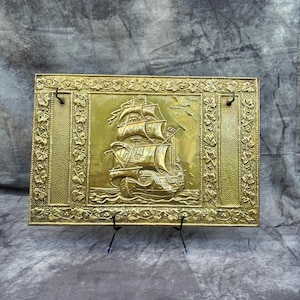May include: A rectangular brass wall hanging featuring a detailed relief of a sailing ship at sea. The ship has multiple sails and is surrounded by decorative elements, including a floral border. The brass has a golden hue.