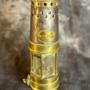 Könnte beinhalten: Antike Bergmannslampe aus Messing mit einem transparenten Glasgehäuse und einem Griff. Die Lampe hat einen zylindrischen Körper mit einer Reihe kleiner Löcher in der Nähe der Oberseite. Die Nummer "3080" ist auf der Vorderseite gestempelt.