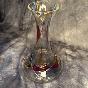 May include: A clear glass vase with a flared top and a rounded base. The vase is decorated with geometric patterns in red, gold, gray, and black. The rim of the vase has a gold trim.