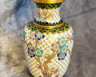 May include: A small, white cloisonné vase with a floral design. The vase has a gold rim and base, and is decorated with blue, pink, and green flowers.