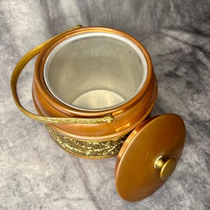 May include: A vintage copper ice bucket with a lid and handle. The bucket has a brass handle and a decorative band around the base. The lid is partially open, revealing the silver interior.