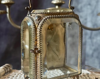 May include: A rectangular carriage clock case featuring beveled glass panels framed by ornate, aged brass metalwork. The structure includes a decorative top handle, intricate filigree corner accents, and four small, detailed feet at the base.