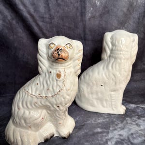Op de afbeelding: Twee witte keramische hondenbeeldjes. De honden zitten rechtop met gedetailleerde gezichtskenmerken, waaronder gele ogen en bruine accenten op de neus en oren. Elke hond heeft een goudkleurige ketting om de nek. De beeldjes hebben een vintage uitstraling.