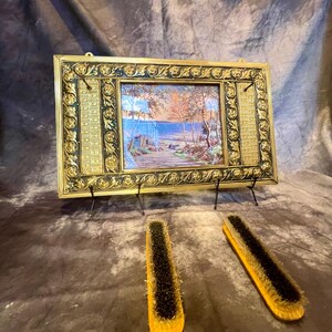 May include: A gold-framed picture with a landscape scene and a black stand. Two wooden shoe brushes with black bristles are in the foreground.