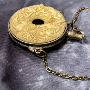 Op de afbeelding: Een vintage, crèmekleurige, ronde metalen hanger met een ketting. De hanger heeft een gedetailleerd, gesneden ontwerp met naakte figuren. De hanger is gesloten en heeft een kleine, ronde opening in het midden.