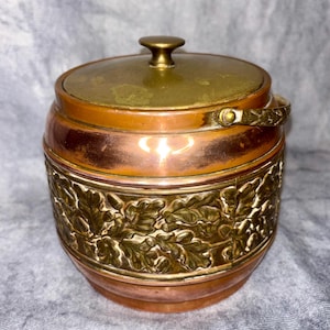 May include: A copper and brass container with a lid and handle. The container features a decorative band of embossed leaves. The lid and handle are brass, while the body is copper. The container is likely vintage.