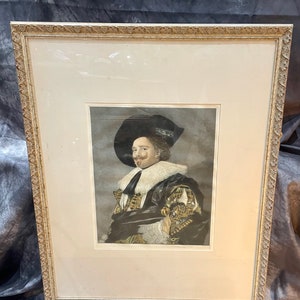 May include: A framed black and white portrait of a man wearing a black hat and a white collar. The man is wearing a dark jacket with gold embroidery. The portrait is set against a gray background.
