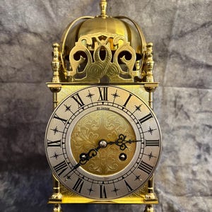 Può includere: Un orologio St. James dorato con una parte superiore a cupola e un quadrante rotondo. Il quadrante dell'orologio presenta numeri romani e dettagli ornati. L'orologio ha un design decorativo ed è sostenuto da piccoli piedini.