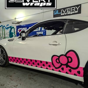 May include: A white car with a pink Hello Kitty bow and heart design on the side. The car is in a garage with a sign that says "9Livery Wraps".