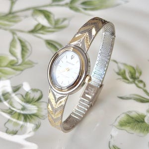 May include: A silver and gold-toned wristwatch with an oval face and a mesh band. The watch face is white with silver hands and a small diamond accent. The band features a leaf design. The watch is displayed on a white surface with green floral patterns.
