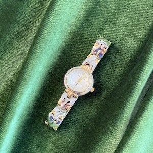 Op de afbeelding: Een zilver- en goudkleurig polshorloge met een witte wijzerplaat en gouden wijzers. De horlogeband heeft een decoratief ontwerp met blauwe en groene accenten. Het horloge ligt op een groene fluwelen achtergrond.