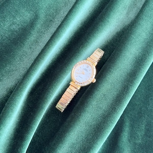 Puede incluir: Un reloj de pulsera dorado con esfera ovalada y blanca. El reloj presenta un bisel adornado con pequeños cristales transparentes y una correa dorada. El reloj se encuentra sobre un fondo de terciopelo verde oscuro.
