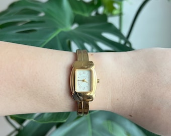 May include: A gold-toned wristwatch with a rectangular face and Roman numeral hour markers. The watch has a gold-toned metal band.