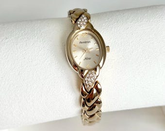 Puede incluir: Reloj Armitron dorado con esfera ovalada y correa trenzada. La esfera es de color claro con la palabra "Now" y la marca Armitron. La correa presenta detalles decorativos de cristal.