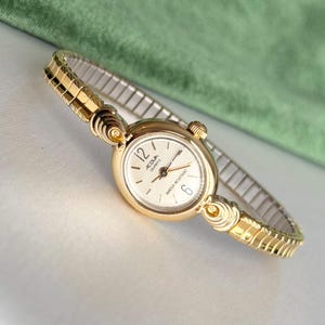 Könnte beinhalten: Goldfarbene Armbanduhr mit cremefarbenem Zifferblatt und schwarzen Ziffern. Die Uhr hat ein flexibles Goldband. Auf dem Zifferblatt steht "ACQUA QUARTZ WATER RESISTANT". Klassisches, elegantes Design.