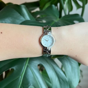 May include: A silver wristwatch with a white face and a decorative band. The watch face has black Roman numeral hour markers and a black second hand. The watch face also has the word "GENEVA" and the word "QUARTZ" printed on it.