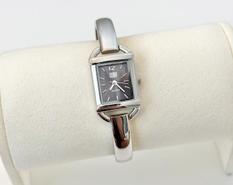 Könnte beinhalten: Eine rechteckige Uhr in Silber mit einem dunkelgrauen Zifferblatt. Die Uhr hat ein silbernes Armband und das Zifferblatt zeigt die Zahl 12 und die Worte "Faded Glory". Die Uhr wird auf einer weißen Oberfläche präsentiert.