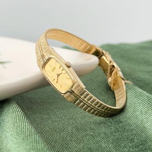 May include: A gold-toned Seiko Quartz wristwatch with a rectangular face and a textured band. The watch features a gold dial with black hands and markers. The band has a textured, woven pattern. The watch is displayed on a white surface against a green background.