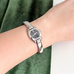 May include: A silver-tone Citizen Quartz watch with a polished bangle-style band and an oval-shaped case. The watch features a black dial with simple silver-tone hands and a small crown on the side, presenting a sleek and minimalist aesthetic.