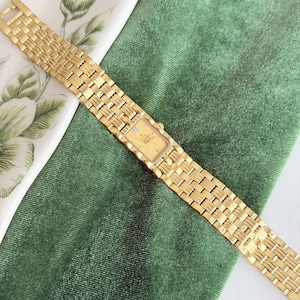 May include: A gold-toned wristwatch with a rectangular face and a woven band. The watch face has a small diamond accent. The watch is displayed on a green velvet surface.