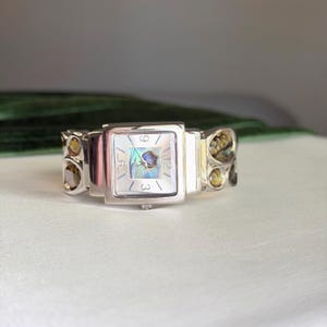 Könnte beinhalten: Eine silberfarbene Armbanduhr mit quadratischem Zifferblatt und einer Einlage aus irisierendem Abalone-Muschelgehäuse. Die Uhr hat silberne Zeiger und Ziffern an den 3-, 6-, 9- und 12-Uhr-Positionen. Das Armband besteht aus silberfarbenen Gliedern mit kreisförmigen Abalone-Akzenten.