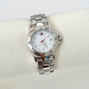 May include: A silver-toned Tommy Hilfiger watch with a white face, featuring black numerals and a date display. The watch has a stainless steel band and a red second hand. The Tommy Hilfiger logo is visible on the watch face.