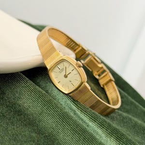 May include: A gold Citizen Quartz watch with a rectangular face and a matching mesh strap. The watch has a cream dial with black hour markers and hands. The watch is displayed on a green velvet surface.