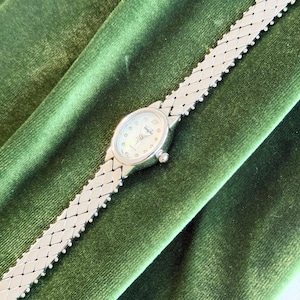 Puede incluir: Un reloj de pulsera plateado con una pulsera de eslabones rectangulares y una esfera redonda. La esfera del reloj tiene un aspecto nacarado con pequeños marcadores de hora y agujas finas. El reloj se muestra sobre una superficie de terciopelo verde.