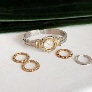 May include: A silver-tone cuff watch with a round white face and gold-tone accents. The watch features a rope-textured bezel and includes four interchangeable rings in gold and silver tones, some embellished with small colorful stones.