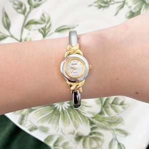 May include: A silver and gold-toned quartz watch with a white face and the brand name "GITANOS". The watch is a bangle style, with a silver band and gold accents. The watch is on a wrist.