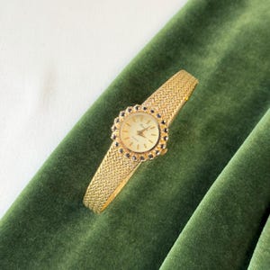 May include: A gold-toned vintage wristwatch with a woven band and a round face. The watch face is surrounded by a decorative bezel with dark blue accents. The watch is displayed on a dark green velvet surface.