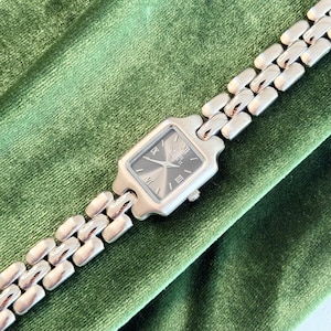 Puede incluir: Reloj de pulsera plateado con esfera rectangular y correa de eslabones. La esfera del reloj es gris oscuro con números romanos y agujas plateadas. El reloj se muestra sobre una superficie de terciopelo verde.