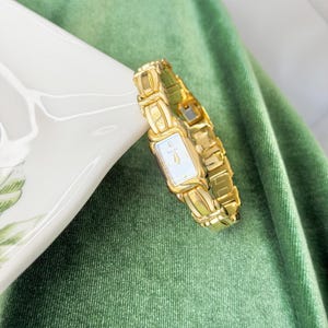 Puede incluir: Un reloj de pulsera dorado con una esfera rectangular y una pulsera de eslabones. La esfera del reloj es blanca con agujas y marcadores dorados. El reloj se muestra sobre una superficie blanca sobre un fondo verde.