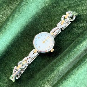 Könnte beinhalten: Silber- und goldfarbene Armbanduhr von Anne Klein mit weißem Zifferblatt. Das Armband ist im Kettendesign mit goldenen Akzenten. Das Zifferblatt zeigt das Anne Klein Logo und die Zahlen 12, 3, 6 und 9.