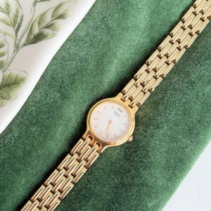 May include: A gold Seiko quartz watch with a white face and gold hands. The watch has a gold link strap and is set against a green velvet background. The watch face has small gold markers and the Seiko logo.