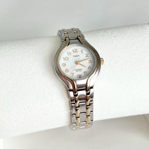 May include: A silver and gold-tone Timex watch with a white face. The watch features gold-tone hands and hour markers, with the numbers 1 through 12. The watch band is a combination of silver and gold-tone links. The watch face includes the text "TIMEX" and "INDIGLO WR 30M".
