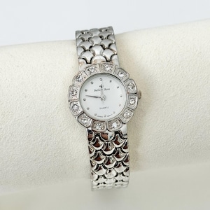 May include: A silver-toned Bella Rose quartz watch with a white face and a decorative bezel set with clear crystals. The watch band has a textured, scale-like design. The watch is displayed on a white surface.