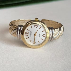May include: A round analog wristwatch with a gold-tone bezel and a white face featuring the text STUDIO and QUARTZ. The dial has gold-tone hands and numerals at 3, 6, and 12. The cuff-style band features alternating gold and silver textured links.