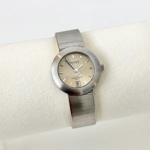 May include: A silver-toned wristwatch with a round face and a mesh band. The watch face is a light champagne color with silver hands and markers. The watch has the text "Serenger Steel" and "8 ATM Water Resistant Quartz" on the face. The date is displayed at the 6 o'clock position.