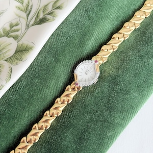 May include: A gold wristwatch with a white face and diamond-studded bezel. The watch has a gold band with a woven design. Two small red gemstones are on either side of the watch face. The watch is displayed on a green velvet surface.