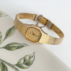Puede incluir: Un reloj Seiko Quartz dorado con una esfera rectangular y una correa de malla. La esfera del reloj tiene un acabado dorado con marcadores de hora y agujas negras. El reloj se muestra sobre una superficie blanca con patrones de hojas verdes.